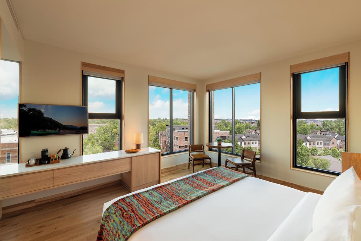 A hotel room with a bed, a desk, a chair, a TV, and large windows offering a city view.
