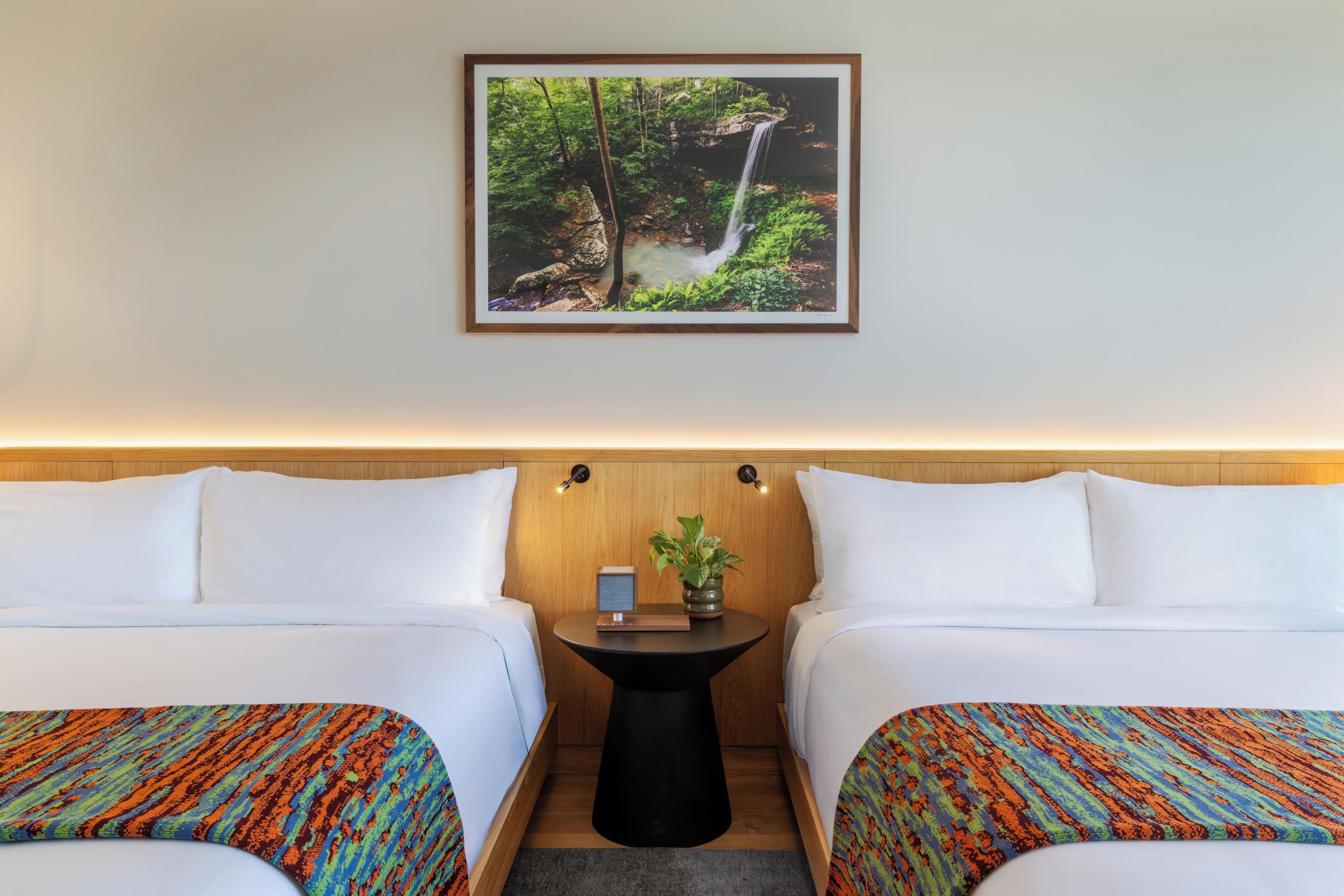 Habitación de hotel con dos camas, una mesita redonda con una planta y un cuadro con una cascada en la pared.
