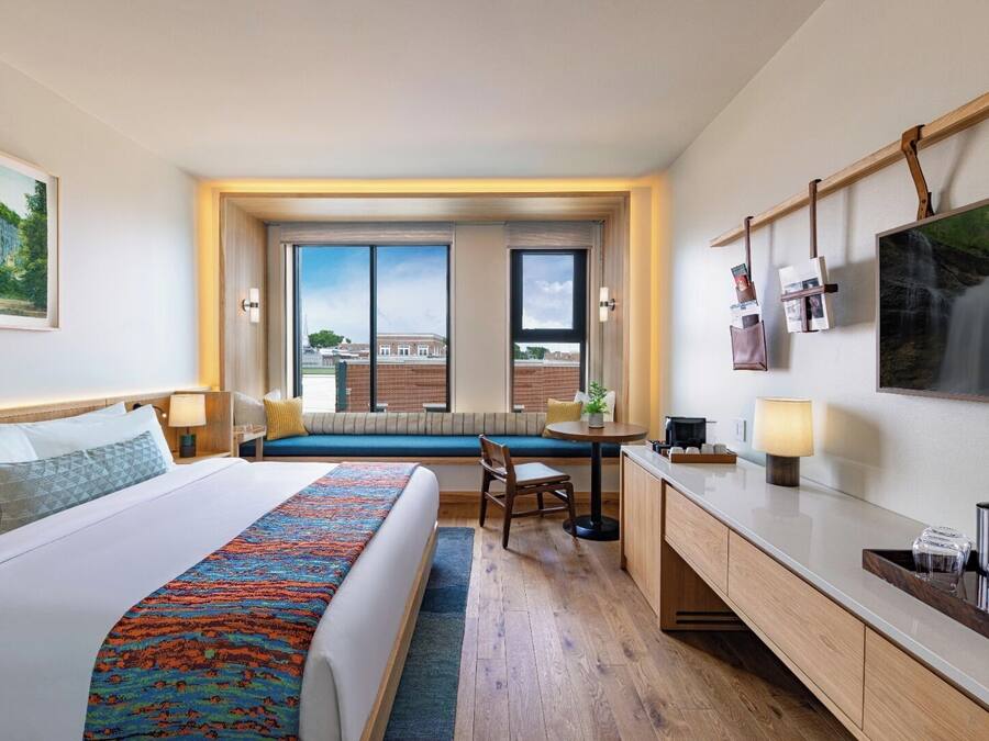 A hotel room with a large bed, a desk, a chair, a window with a view of a building, and a mounted TV.