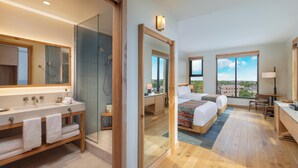 A modern hotel room with a large bed, a desk, a bathroom with a large mirror, and a view of the city. - The Compton (Bentonville)