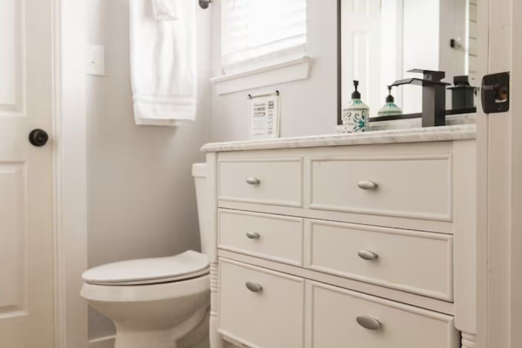 A white bathroom with a toilet, a vanity with drawers, a mirror, and a window with a towel hanging on the wall.
