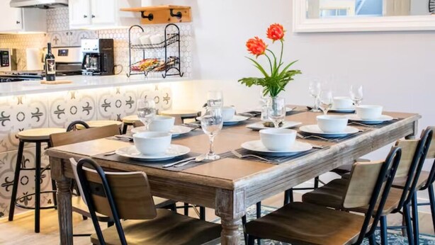 On aperçoit une salle à manger avec une table en bois dressée pour six personnes, entourée de chaises. Au fond, on distingue une cuisine équipée d’appareils modernes et dotée d’une étagère suspendue.
