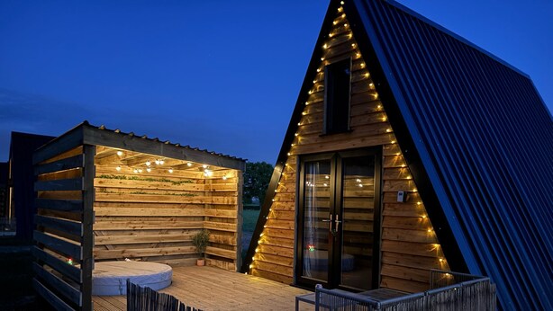 Una cabaña de madera con jacuzzi y luces colgantes, rodeada por una terraza y valla de madera.