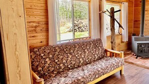 A wooden cabin interior with a sofa, two windows with curtains, a fireplace, and a rustic wooden floor.