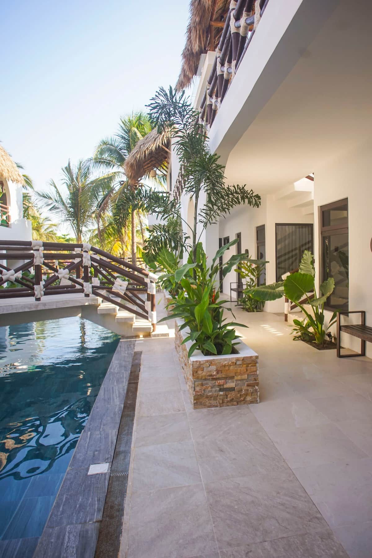 Zona de piscina con vista despejada al agua, rodeada por un edificio blanco con balcón y palmeras.