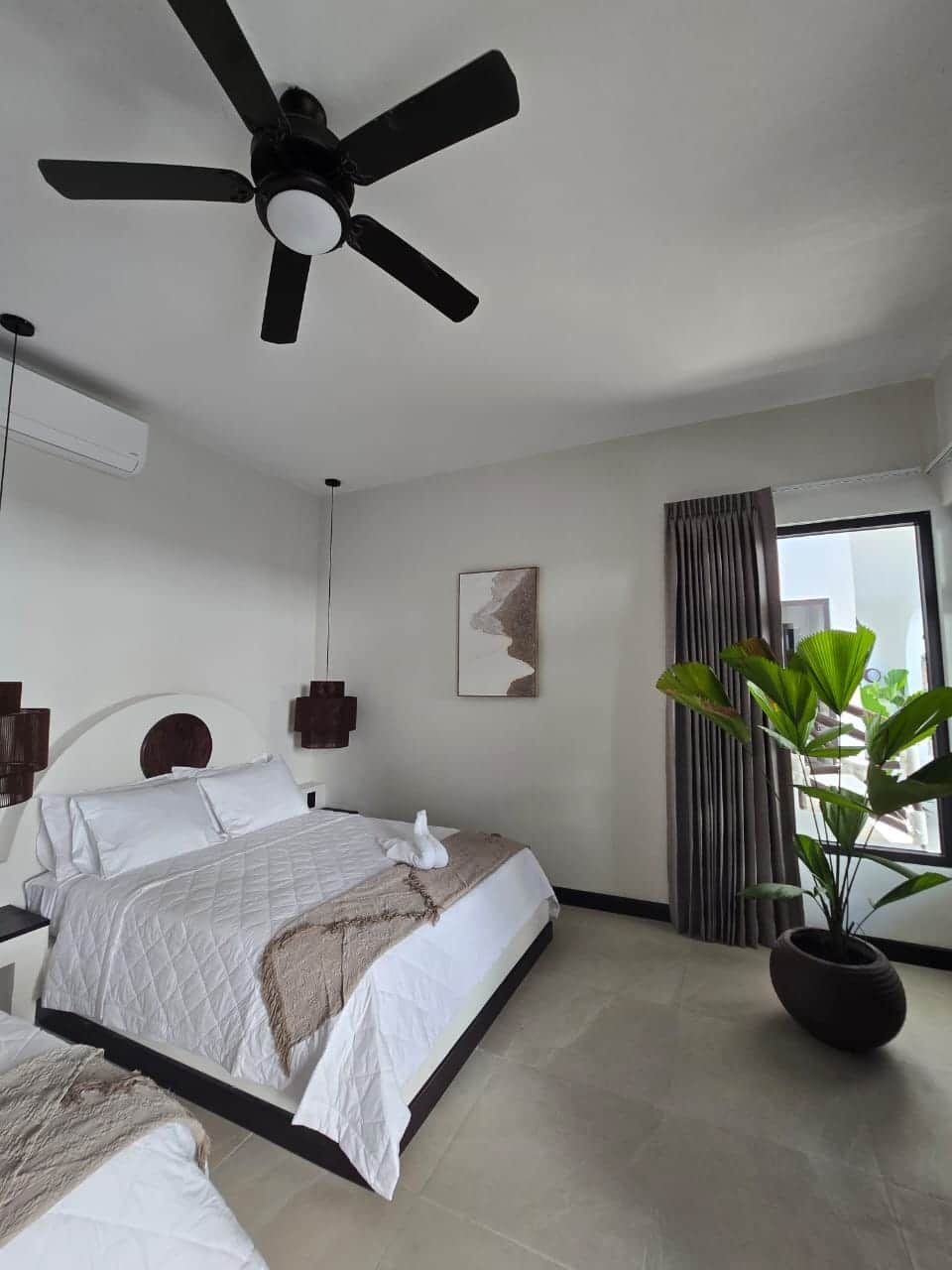 Een hotelkamer met een bed, plafondventilator, kunstwerk, gordijnen en een potplant.