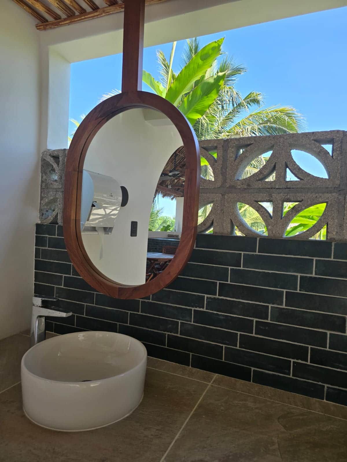 A bathroom with a round mirror, a white sink, and a view of palm trees through a window.