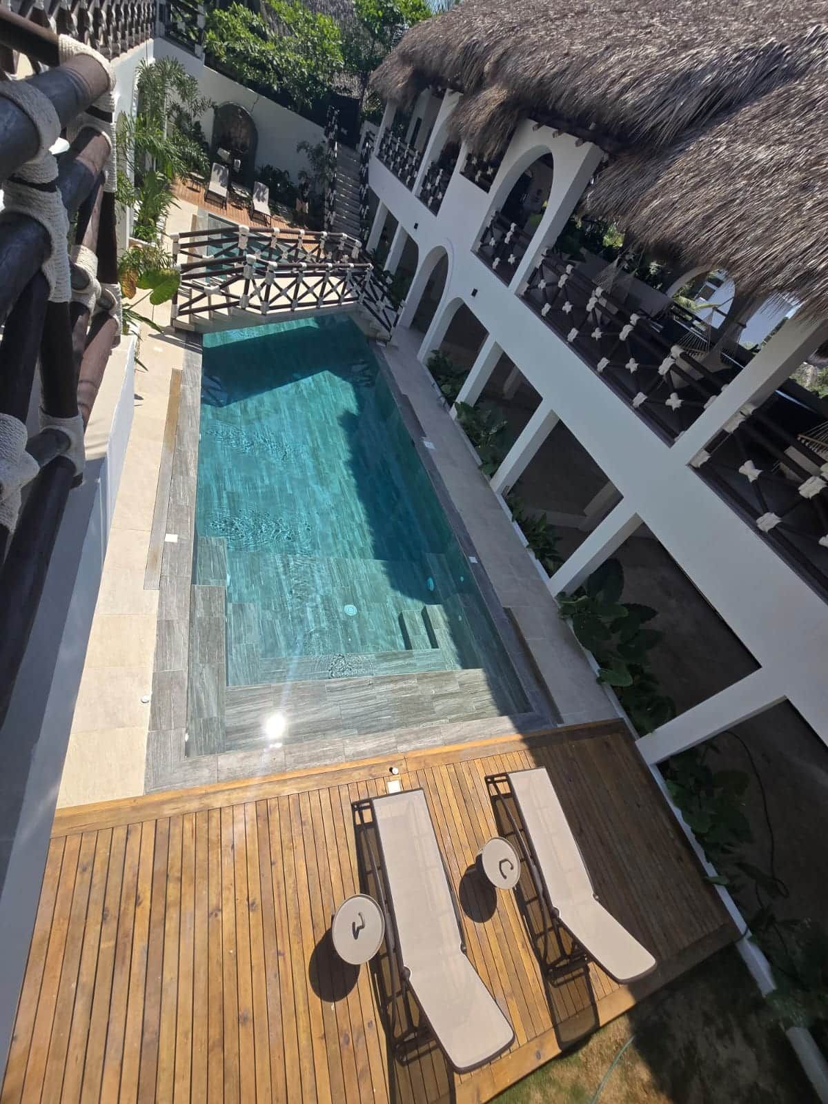 A swimming pool with a wooden deck and lounge chairs, surrounded by white walls and a thatched roof.