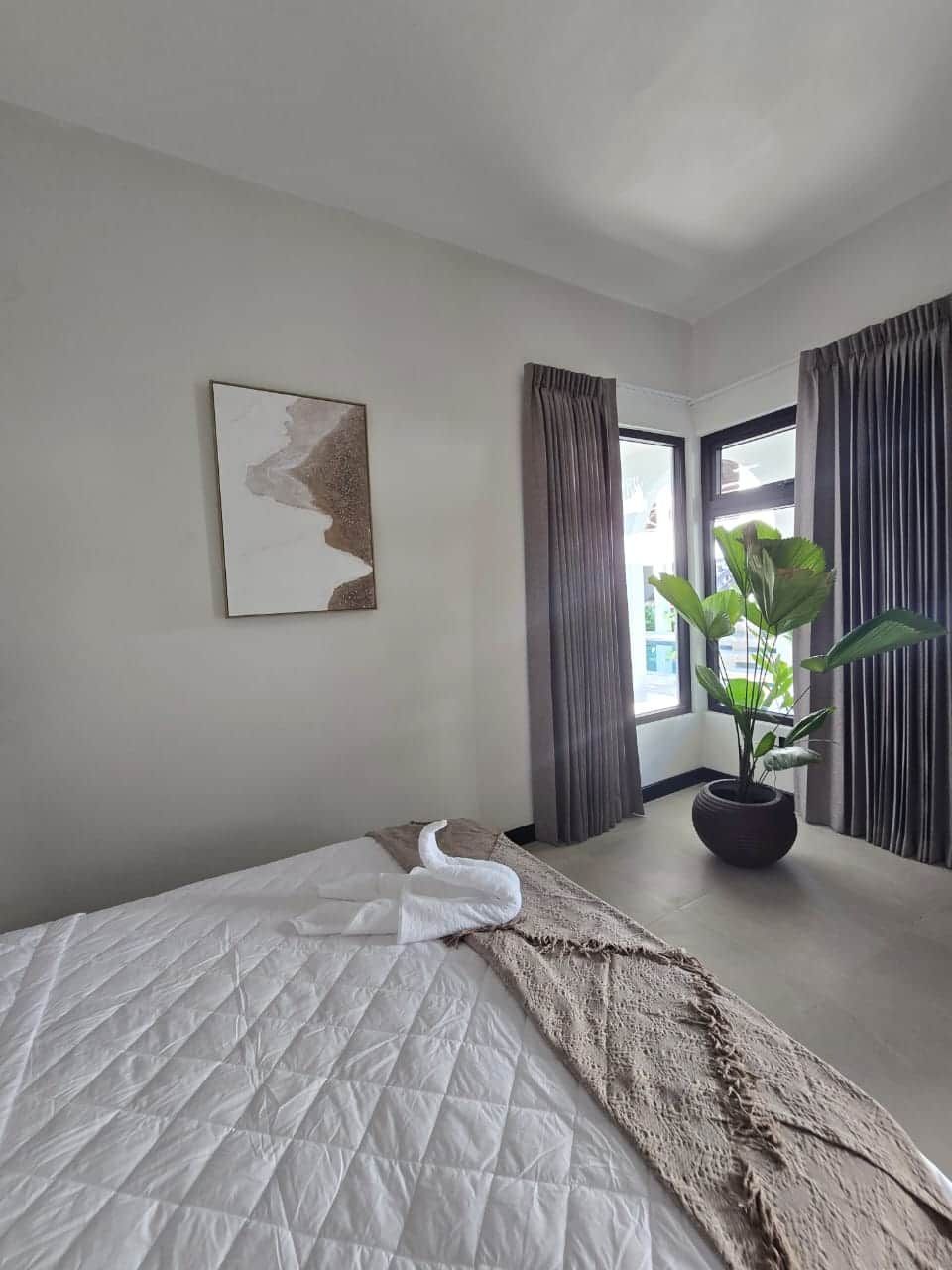 A hotel room with a bed, a potted plant, a painting on the wall, and a window with curtains.