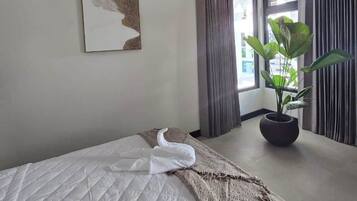 A hotel room with a bed, a potted plant, a painting on the wall, and a window with curtains.