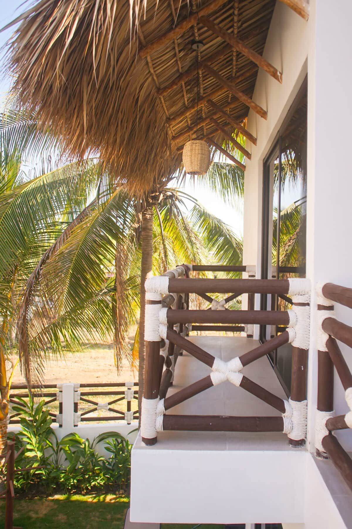 A thatched roof structure with a woven basket hanging from the ceiling, a white railing, and a view of palm trees.