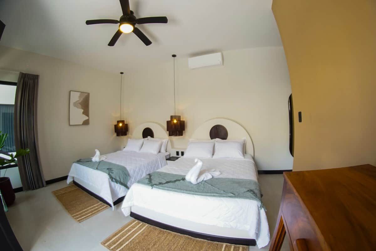 A hotel room with two beds, a ceiling fan, wall-mounted lights, and a wooden nightstand.