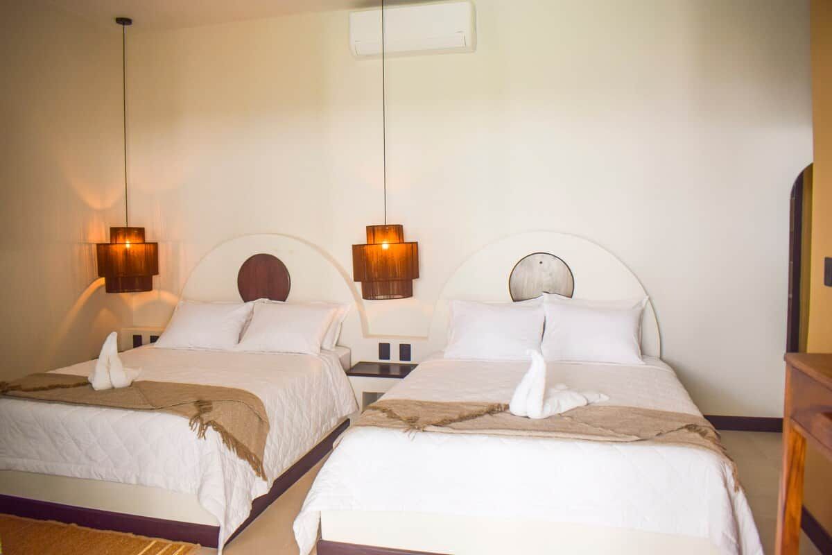 Habitación de hotel con dos camas, cada una con cabecera, mesita de noche y lámpara colgante.