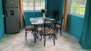 A compact kitchen with a refrigerator, a table with chairs, and a window with a view of greenery. - Jude Apartments - 3-bedroom apartment with AC in  Soufrière (Soufrière)