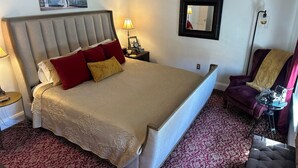 A hotel room with a large bed, a nightstand with a lamp, a chair, a small table with a camera, and a framed picture on the wall.
