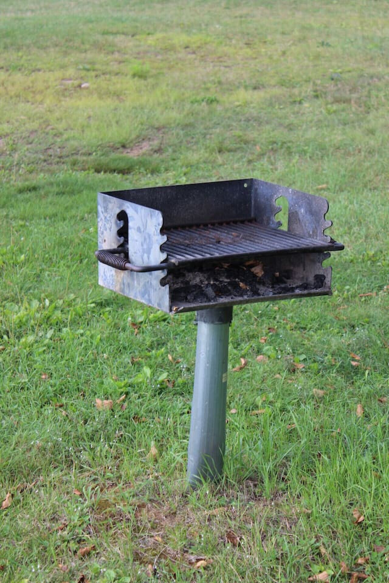 Una barbacoa metàl·lica amb una reixa rectangular i un mànec, subjecta a un pal metàl·lic, situada en un camp amb herba.