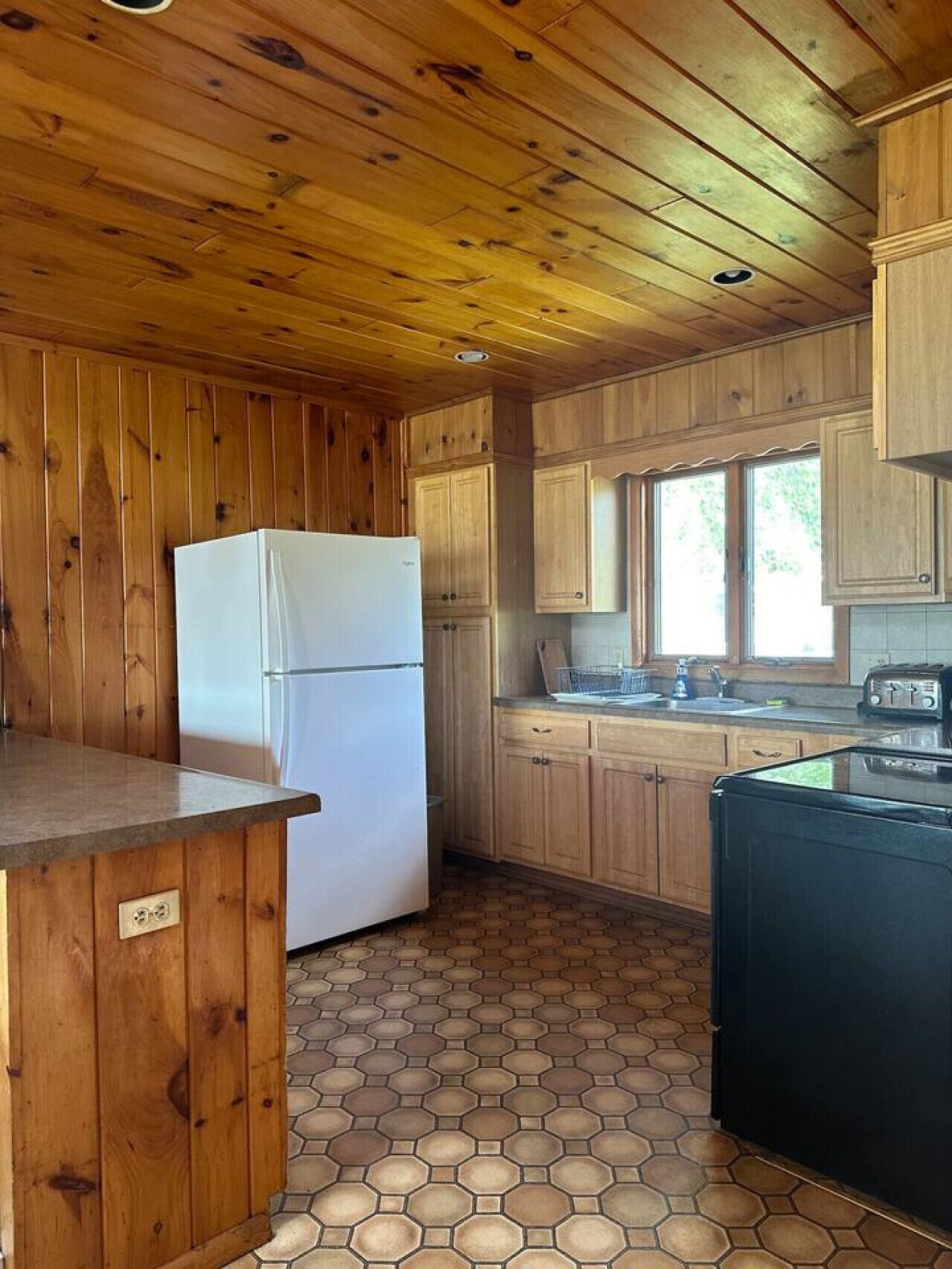 A cabin-style kitchen with wooden walls and floors, a refrigerator, a sink, and a toaster.