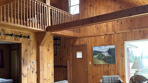 A spacious cabin interior with a wooden ceiling, a loft area with a balcony, a plaid sofa, a wooden rocking chair, and a large window offering a view of the outdoors. - Lakefront-Family & Pet Friendly-Adirondack Cabin (Indian Lake)