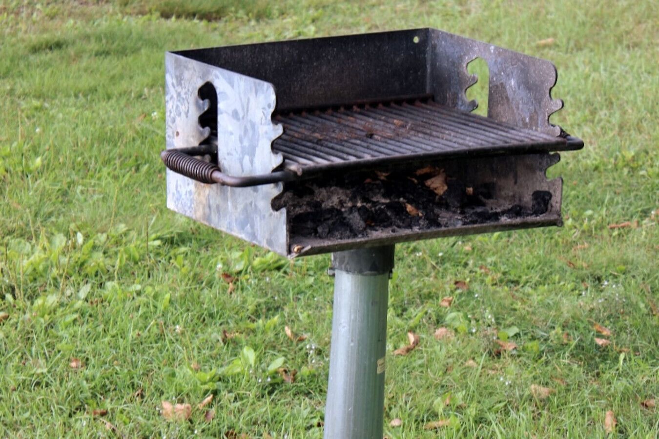 Una parrilla con rejilla metálica y un asa, sobre una superficie cubierta de césped.