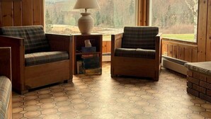 A cozy cabin interior with wooden walls and a large window offering a view of a forest. There are two plaid chairs and a table with a lamp. - Lakefront-Family & Pet Friendly-Adirondack Cabin (Indian Lake)