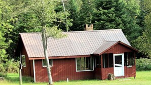Eine kleine, eingeschossige Holzhütte mit einem Metalldach, umgeben von einer grasbewachsenen Wiese und dichtem Baumbestand.