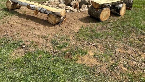 Ein Lagerfeuer, umgeben von Holzstämmen, in einer offenen, grasbewachsenen Gegend mit Bäumen und Bergen im Hintergrund.