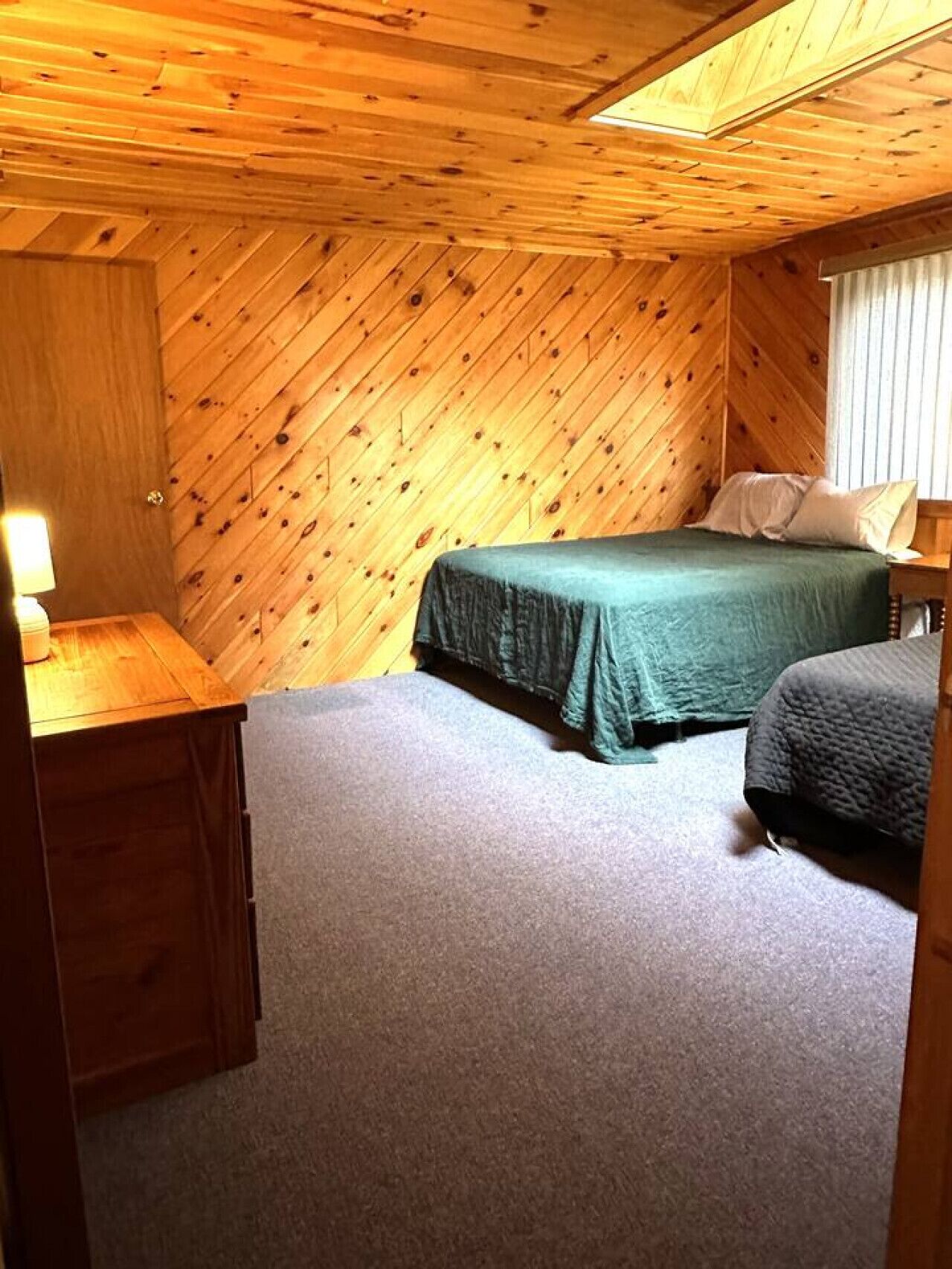 A small, cozy room with a wooden hipped roof ceiling, a single bed with a green bedspread, a small desk with a lamp, and a window with blinds.