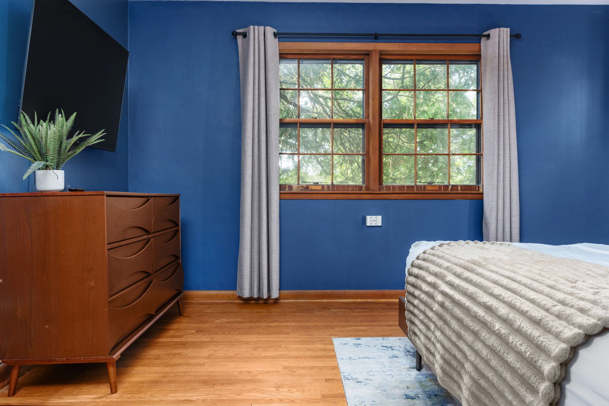 Un dormitorio con un aparador de madera, una cama con manta blanca, una ventana con cortinas y una planta en maceta.