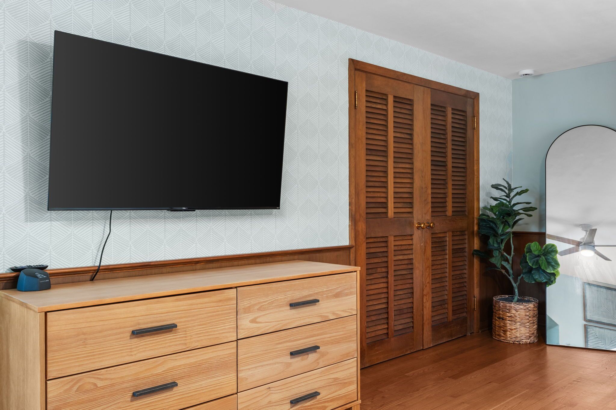 Una habitación con un televisor de pantalla plana montado en la pared, un mueble de madera con cajones, una puerta de madera y una planta en maceta.