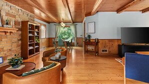 A cozy living room with wooden floors, a brick wall, a bookshelf, a dining area with chairs, and a flat-screen TV.