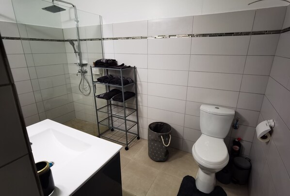 A bathroom with a shower, toilet, and a shelf with folded clothes. - Bas de Villa en campagne (Le Lamentin)