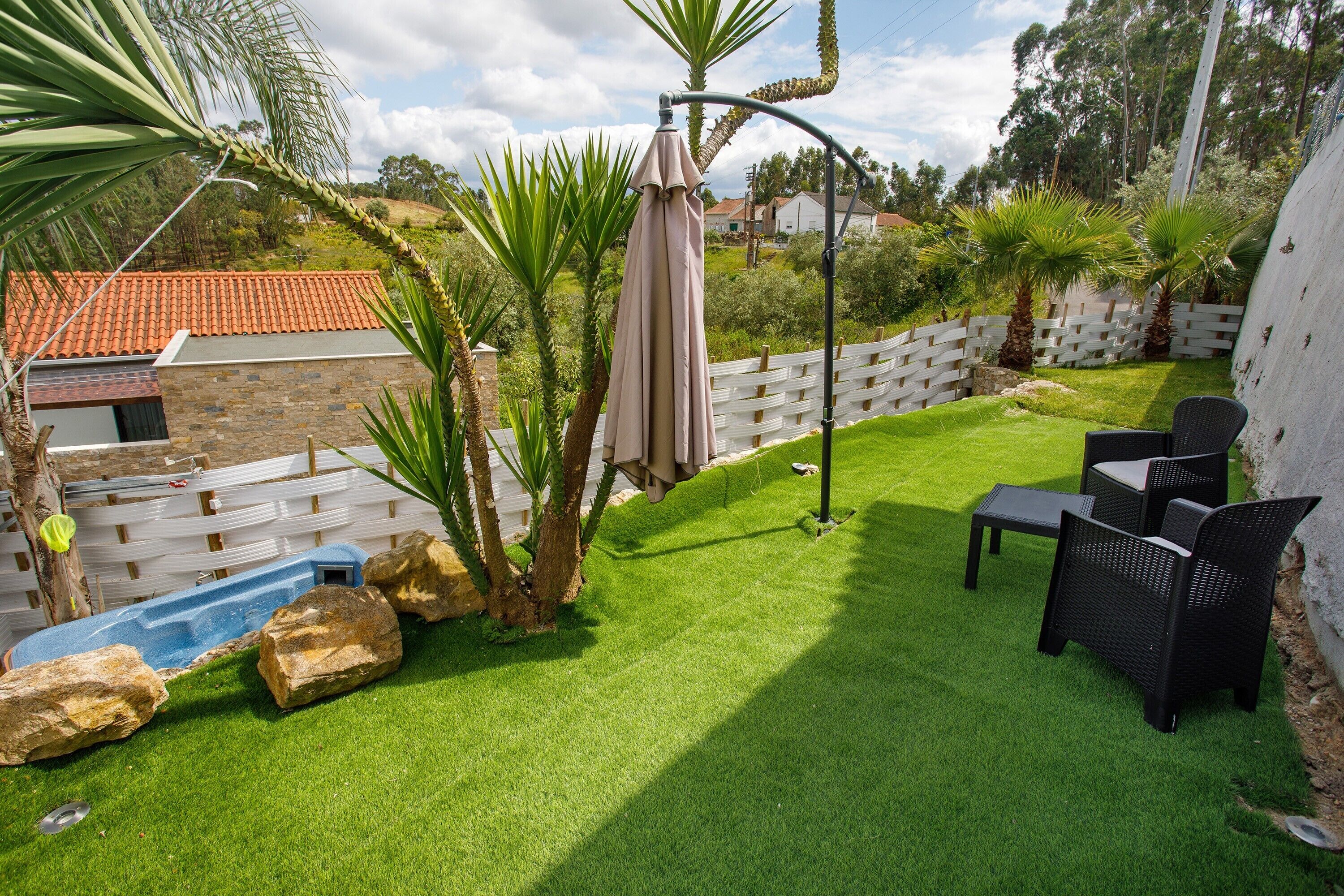 A backyard with artificial grass, a patio table and chairs, a white fence, and palm trees.