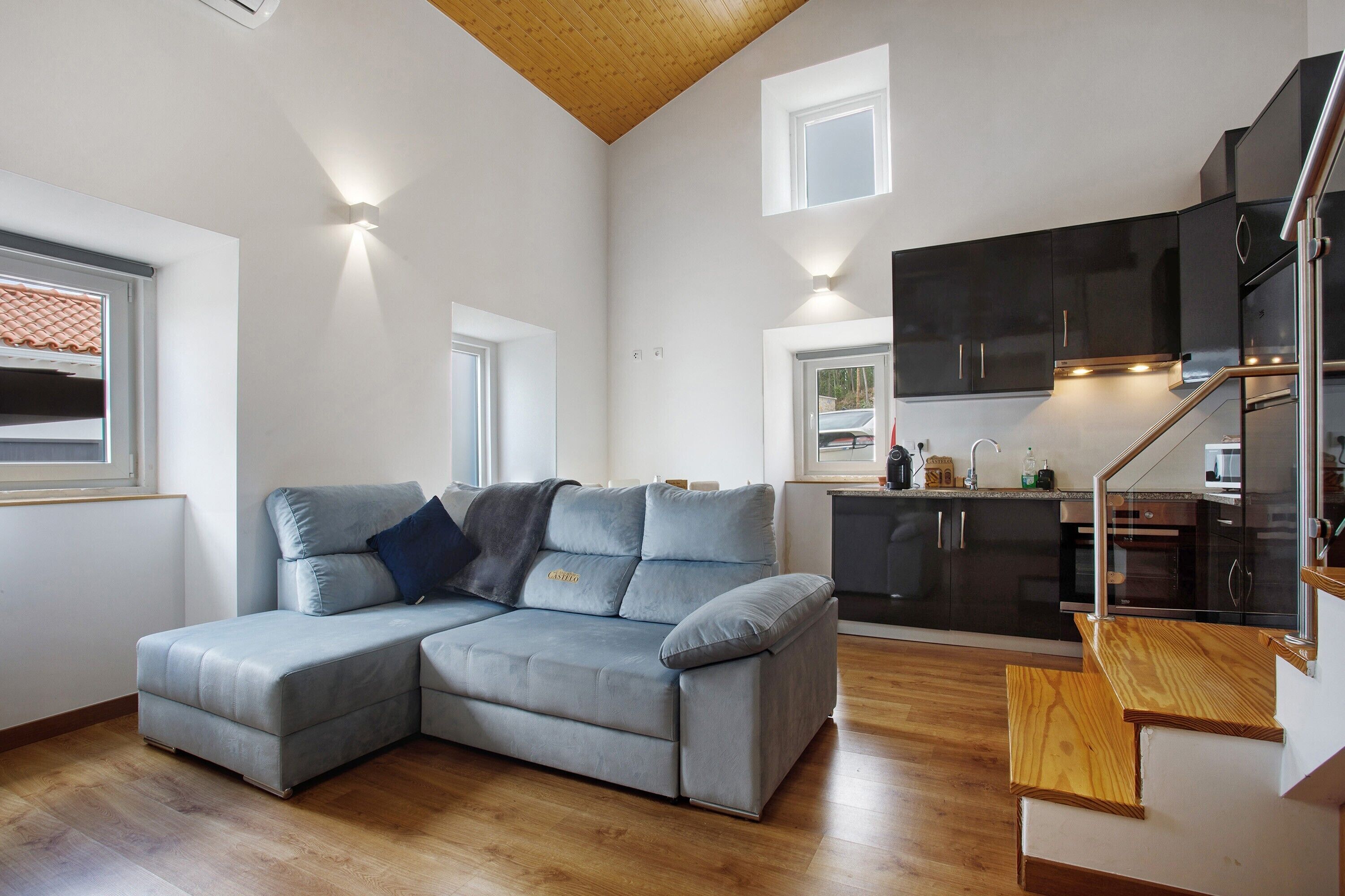 Un espace de vie moderne comprenant un canapé d’angle, un plancher en bois, une cuisine avec des meubles sombres et un escalier avec des marches en bois.