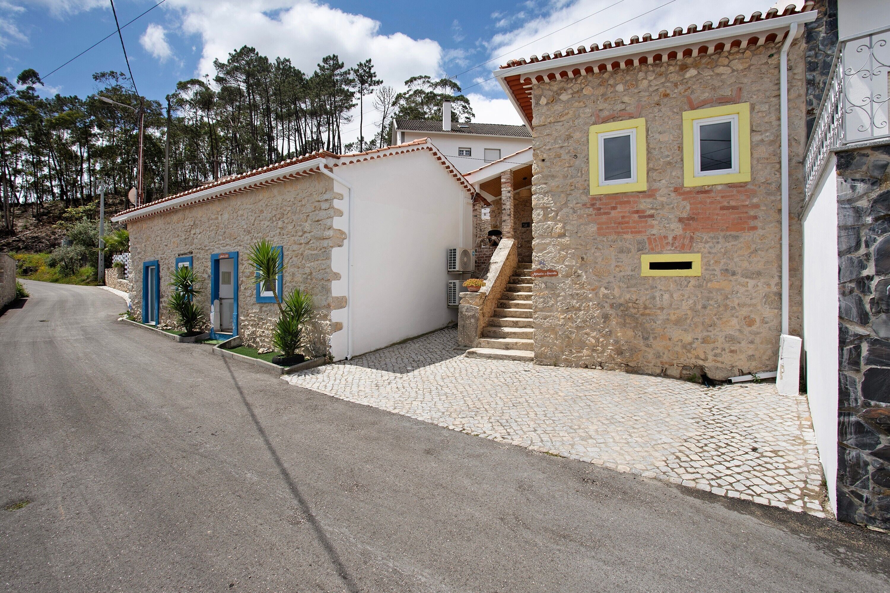 A stone building with a staircase leading to a door, a paved driveway, and a road in the background.
