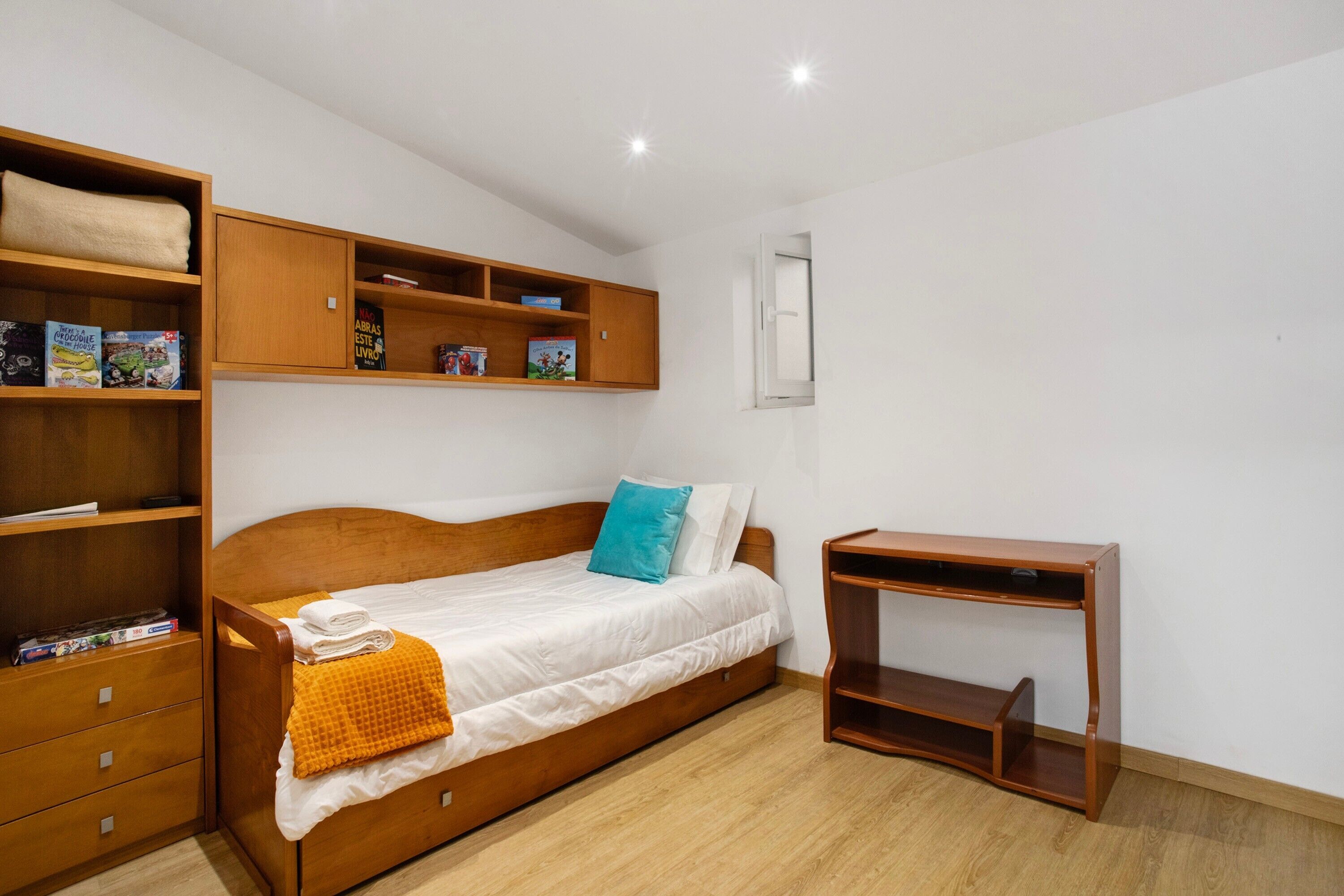 A bedroom with a wooden bed, a nightstand, a shelf with books, and a window.