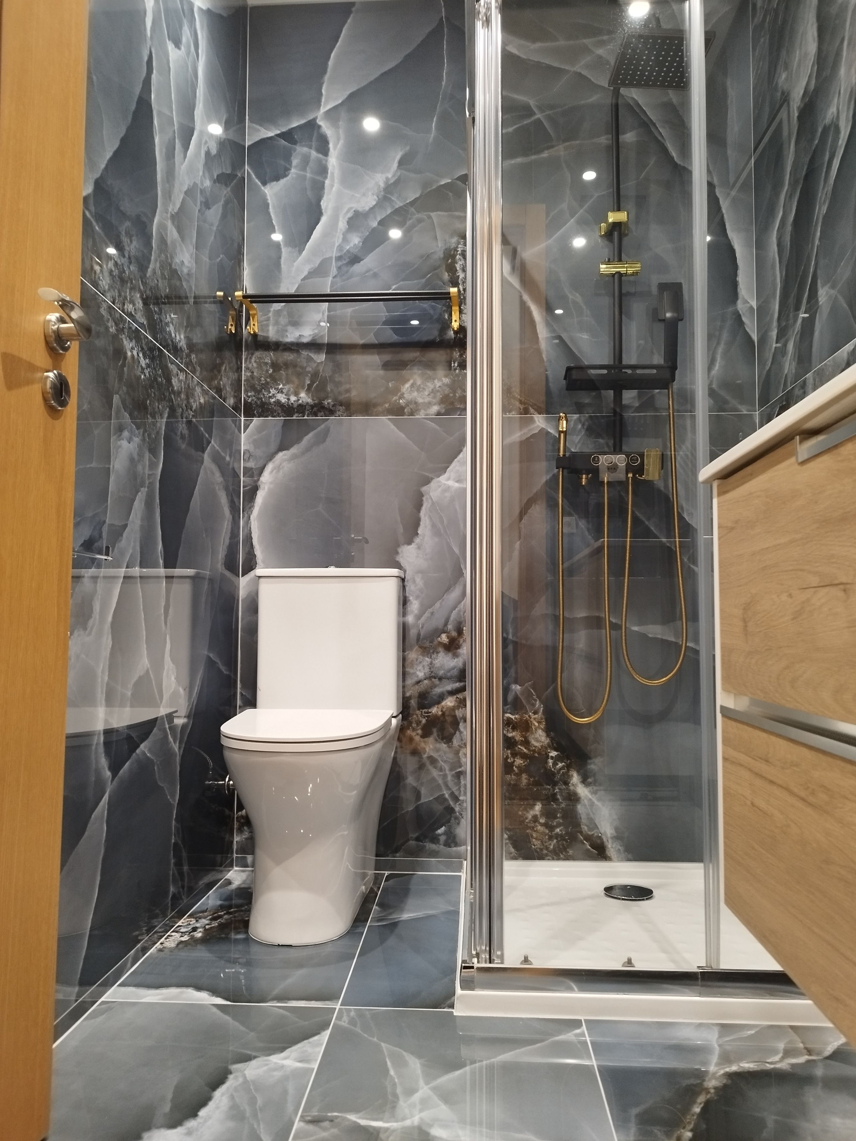 Et badeværelse med marmorgulv, toilet, brusekabine med glasvægge og en håndholdt brusearm, samt en trækommode.