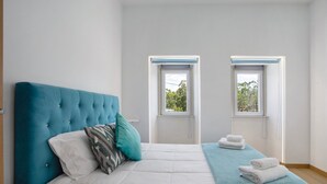 Une chambre avec un grand lit, deux fenêtres et une vue sur les arbres à l’extérieur.