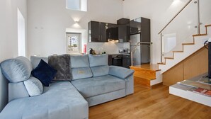 Un espace de vie moderne comprenant un canapé d’angle, une cuisine avec des meubles de couleur sombre et un escalier en bois.