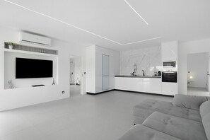 Un salon moderne doté d’un téléviseur à écran plat, d’un canapé gris et d’un coin cuisine avec des meubles blancs.