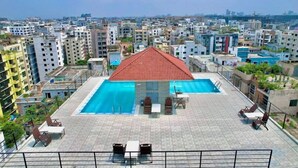 A rooftop swimming pool with a red roof, surrounded by lounge chairs and tables, overlooking a densely built urban area. - 250 sqft 2 Studio Apt Pool and Sauna (Dhaka)