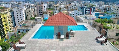 A rooftop swimming pool with a red roof, surrounded by lounge chairs and tables, overlooking a densely built urban area.