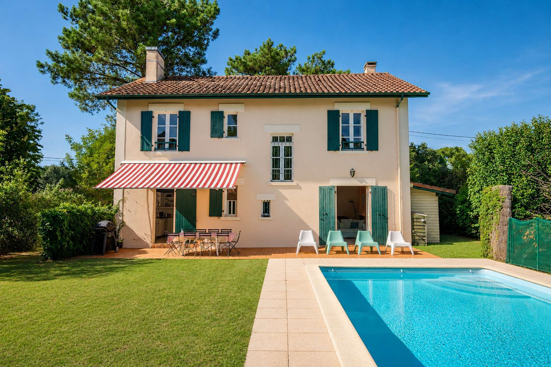 A two-story house with a swimming pool, green shutters, and a red and white striped awning.