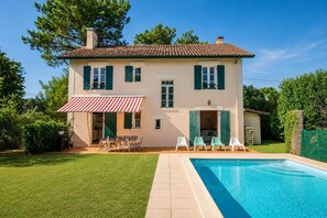 A two-story house with a swimming pool, green shutters, and a red and white striped awning.