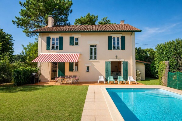 A two-story house with a swimming pool, green shutters, and a red and white striped awning.