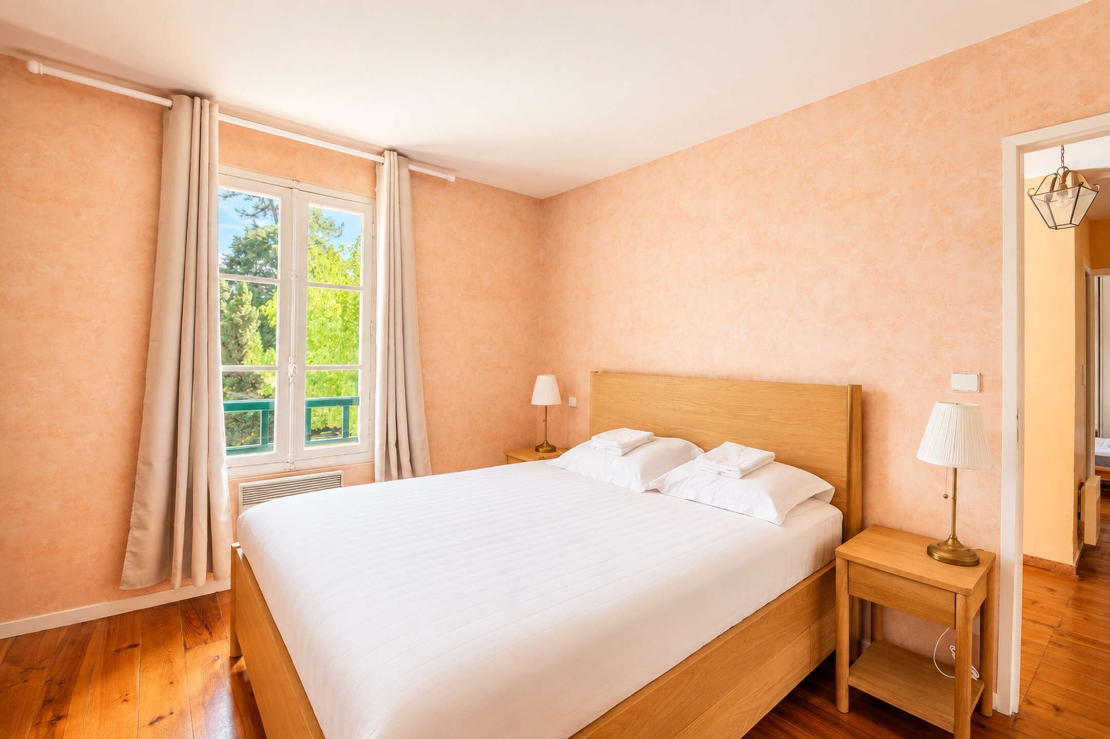 A bedroom with a large bed, two bedside tables, a window with curtains, and a view of greenery outside.