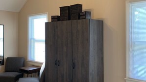 A bedroom with a dark wooden wardrobe, a clock on the wall, a chair, a small table, and a window with blinds.