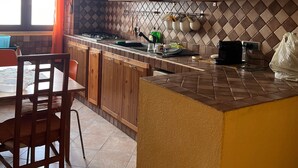 Una cocina con armarios de madera, salpicadero de azulejos, comedor con sillas y ventana con cortinas.