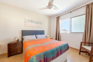Un dormitorio con cama, mesita de noche, silla, ventilador de techo, ventana con cortinas y un cuadro en la pared.