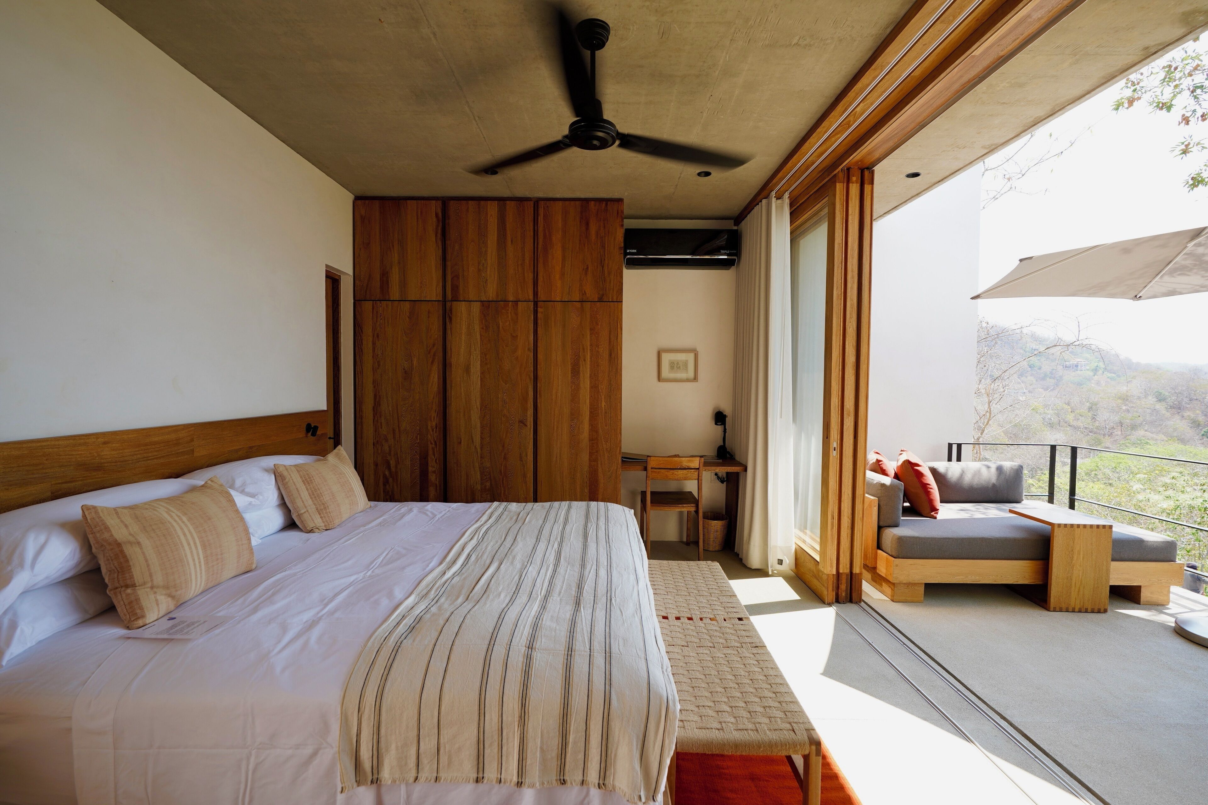 A bedroom with a large bed, a ceiling fan, a wooden wardrobe, a small table, and a balcony with a sofa.