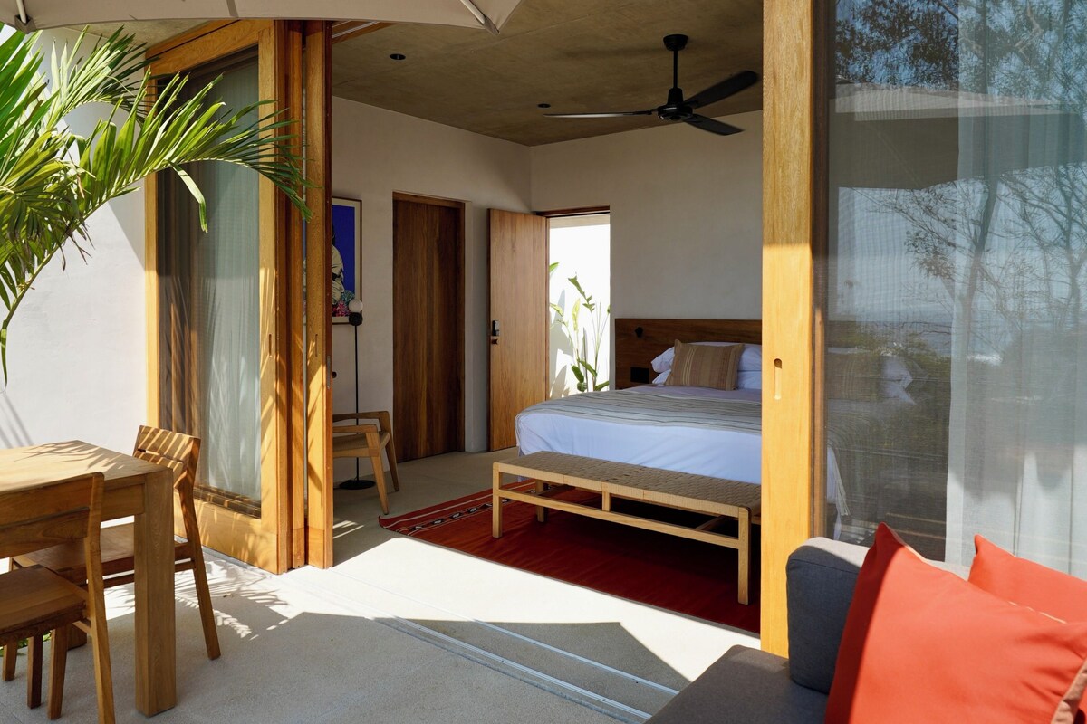 A modern room with a bed, a sofa, a wooden table, and a ceiling fan.